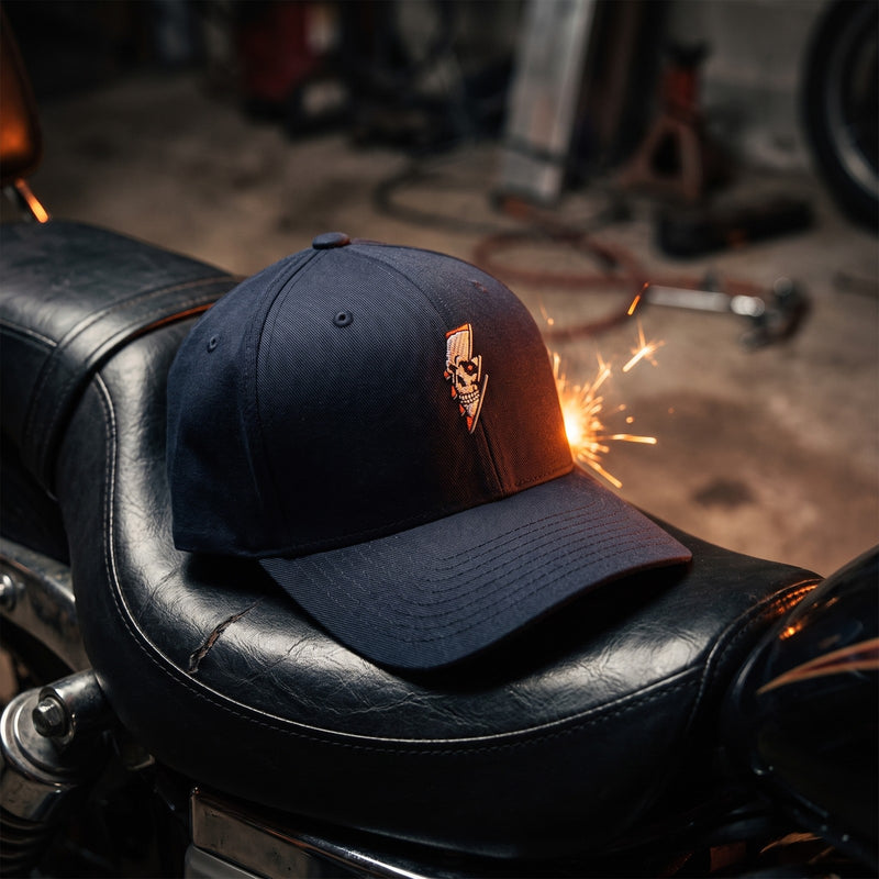 Casquette Flexfit fermée à l'arrière bleu marine brodée d'une tête de mort et d'un éclair posée sur la selle en cuir noir d'une moto dans un garage sombre avec une lueur orange - CHT'M Boutique