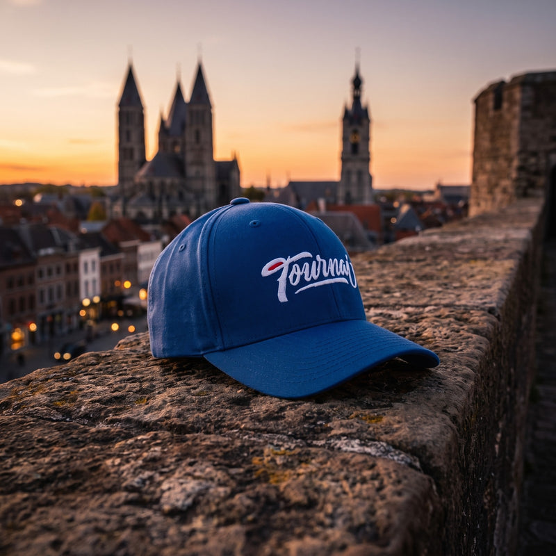 Casquette Flexfit fermée à l'arrière bleu roi brodée Tournai en blanc posée sur un parapet en pierre face à la vieille ville de Tournai et aux tours de la cathédrale au crépuscule - CHT'M Boutique