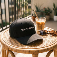 Casquette Flexfit fermée à l'arrière noire brodée TOUT VA BIEN en blanc posée sur une chaise en rotin d'un balcon ensoleillé à côté d'un café glacé et de lunettes de soleil - CHT'M Boutique