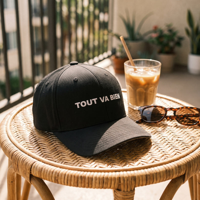 Casquette Flexfit fermée à l'arrière noire brodée TOUT VA BIEN en blanc posée sur une chaise en rotin d'un balcon ensoleillé à côté d'un café glacé et de lunettes de soleil - CHT'M Boutique