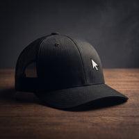 Casquette de geek trucker noire avec curseur de souris brodé, posée sur un plan en bois sombre