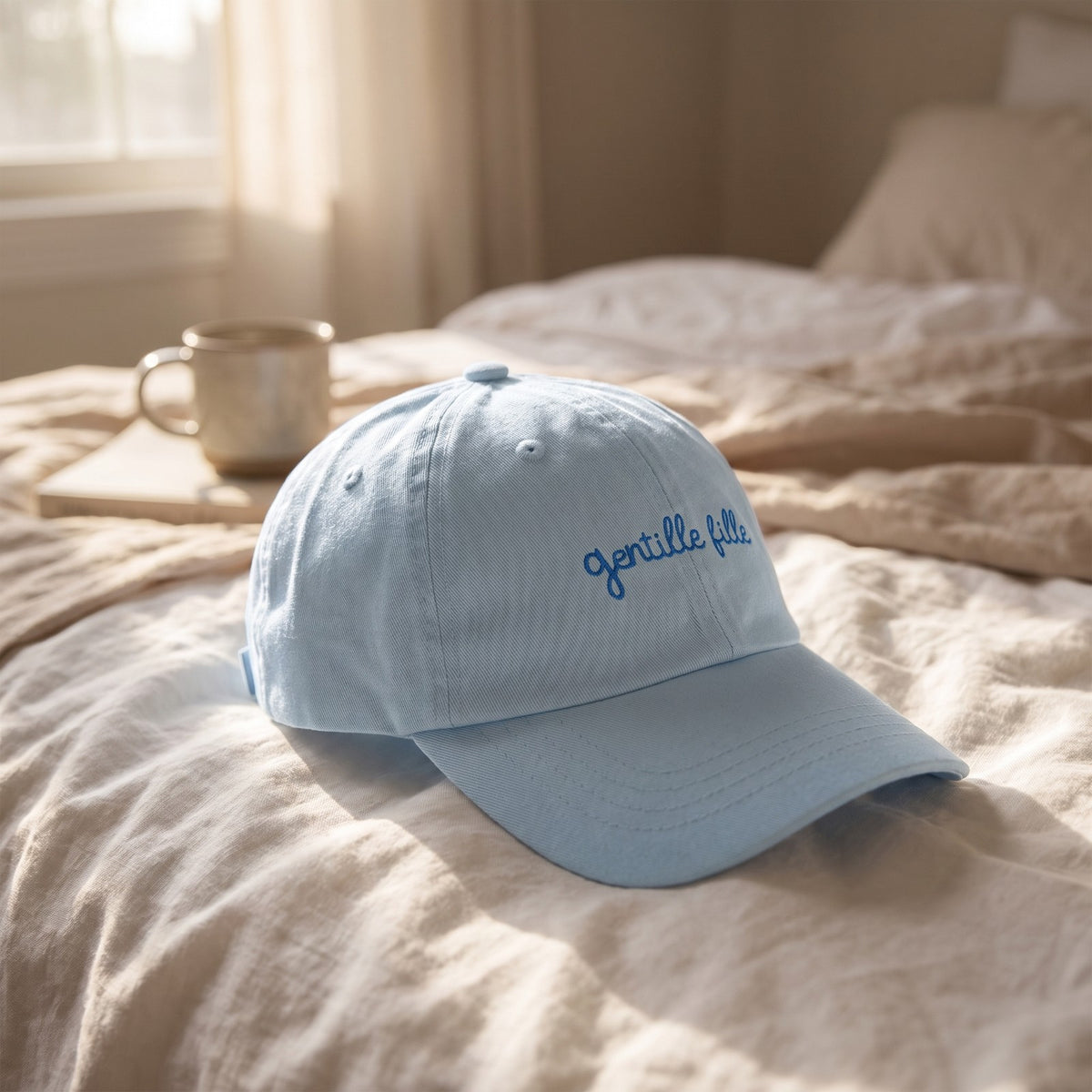 Casquette bleu pastel brodée Gentille Fille posée sur un lit en lin avec un livre et une tasse dans un rayon de soleil - CHT'M Boutique