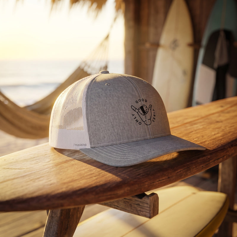 Casquette trucker gris chiné Good Vibes Only avec emblème shaka posée sur un rack de surf dans une cabane de surfeur à l'aube - CHT'M Boutique