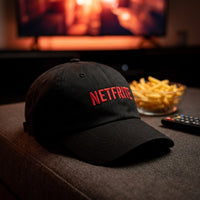 Casquette dad hat noire brodée NETFRITE posée sur un coussin de canapé devant la télé avec un bol de frites - CHT'M Boutique