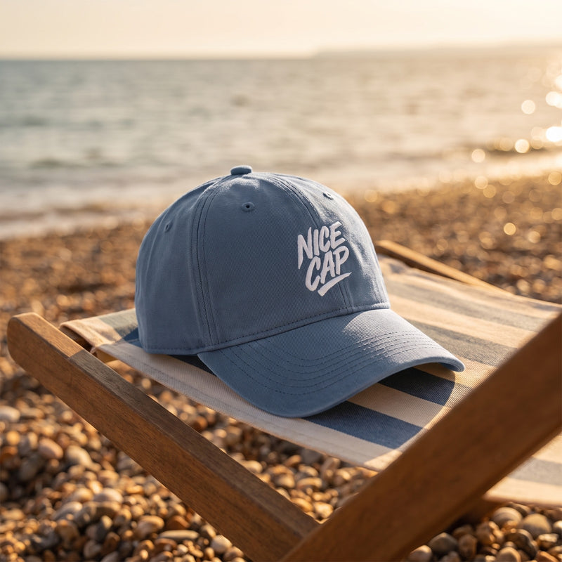 Casquette délavée bleue brodée NICE CAP posée sur un transat rayé sur une plage de galets au coucher du soleil - CHT'M Boutique