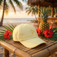 Casquette jaune pastel brodée oiseau du paradis posée sur une table en bambou d'un bar de plage tropical avec un ananas et des hibiscus - CHT'M Boutique
