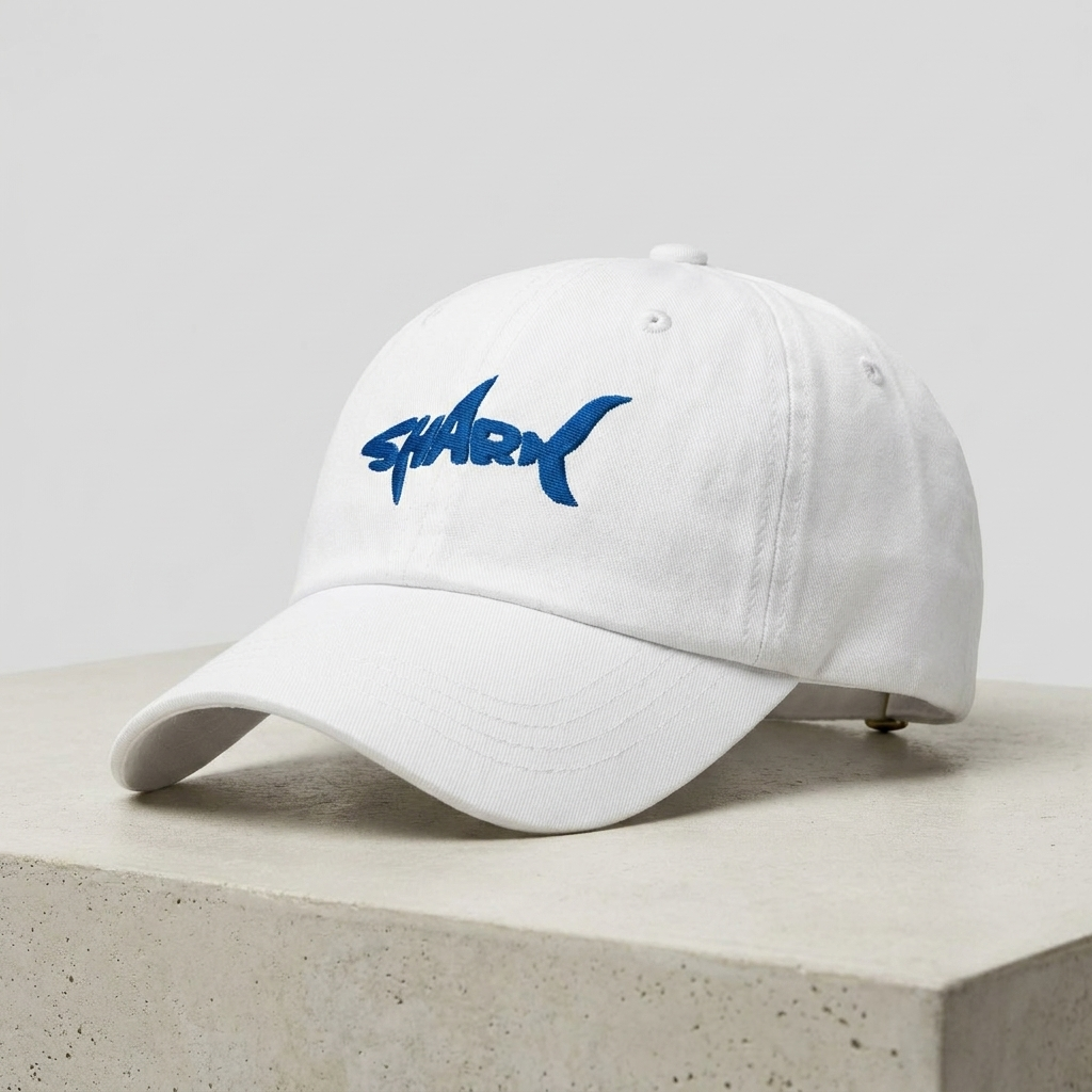 Casquette requin brodée blanche posée sur béton, vue trois-quarts avec broderie SHARK