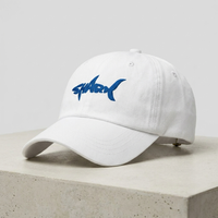 Casquette requin brodée blanche posée sur béton, vue trois-quarts avec broderie SHARK