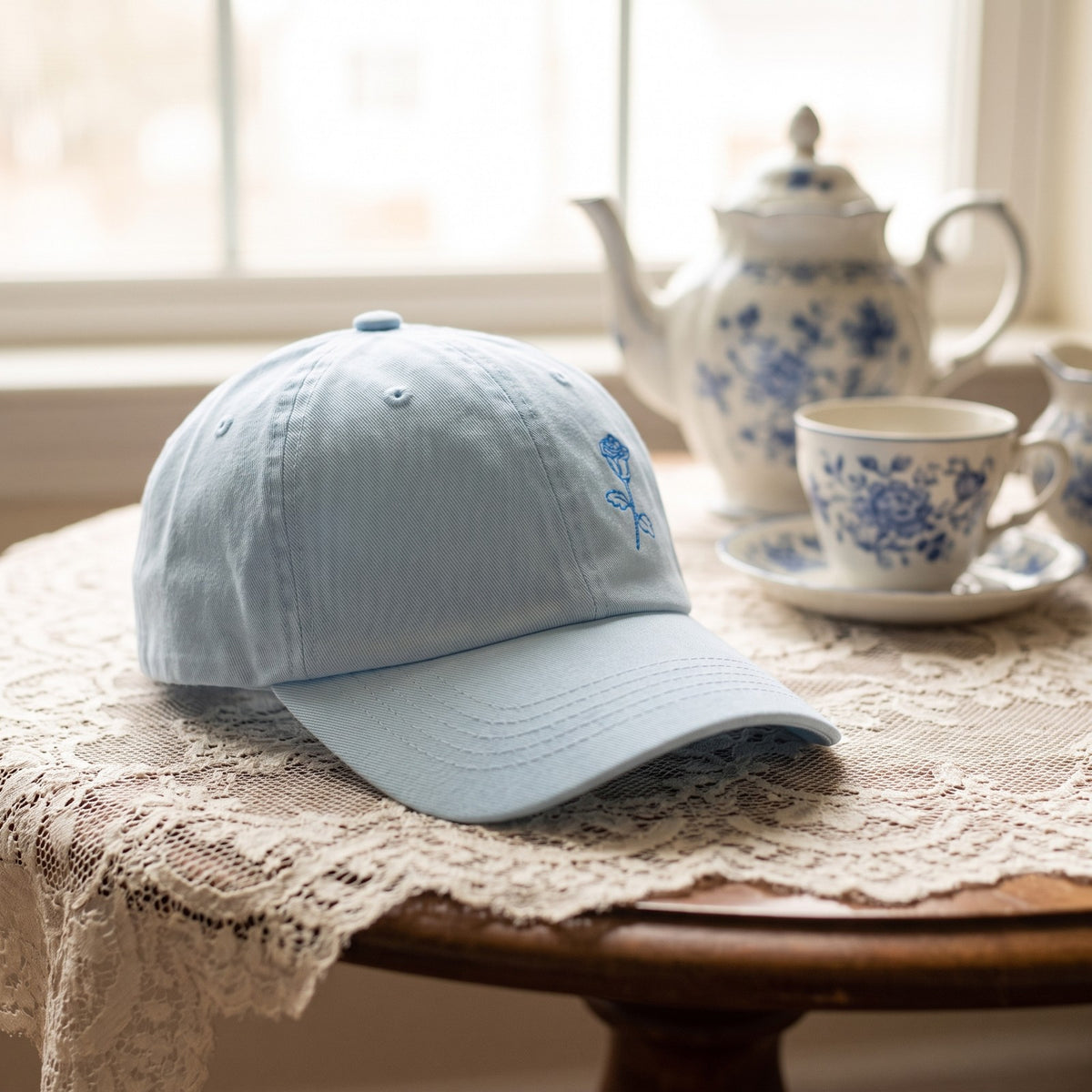 Casquette baseball bleu pastel brodée d'une rose posée sur une table à thé avec un service en porcelaine et une nappe en dentelle - CHT'M Boutique