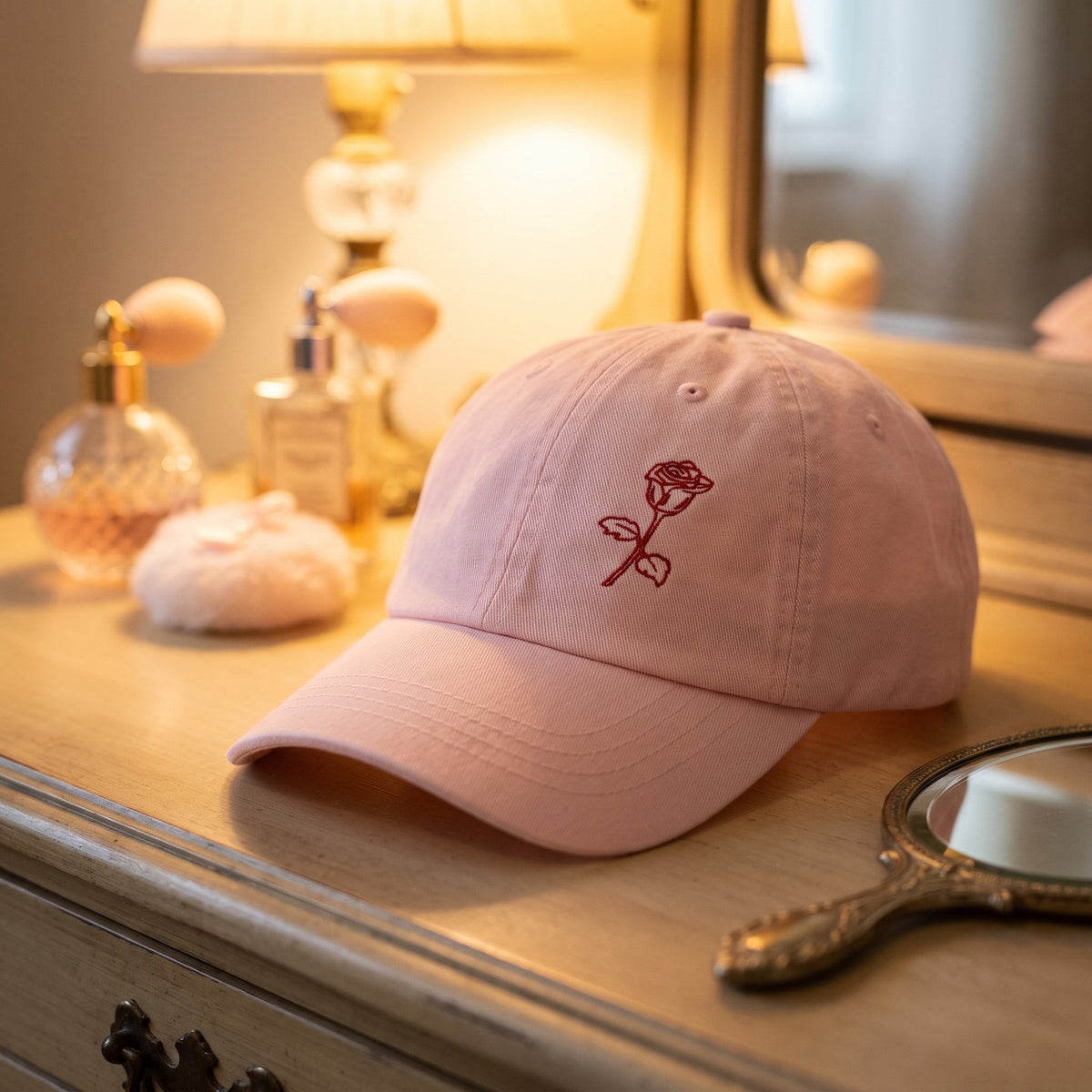 Casquette baseball rose pastel brodée d'une rose posée sur une coiffeuse vintage avec des flacons de parfum - CHT'M Boutique