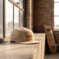 Casquette skate 5 panneaux beige brodée Skate rouge posée sur le rebord d'une fenêtre de loft industriel avec un skateboard - CHT'M Boutique