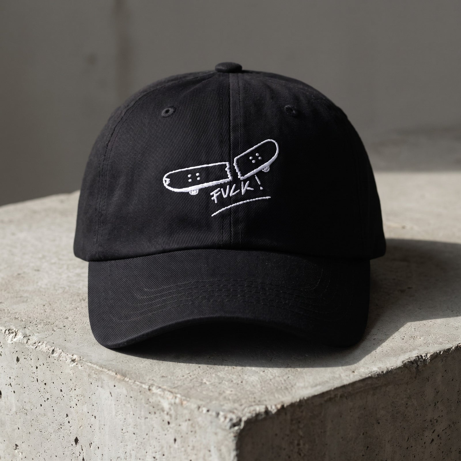 Casquette skate brodée FVCK noire posée sur béton, design skateboard cassé complet vu de face