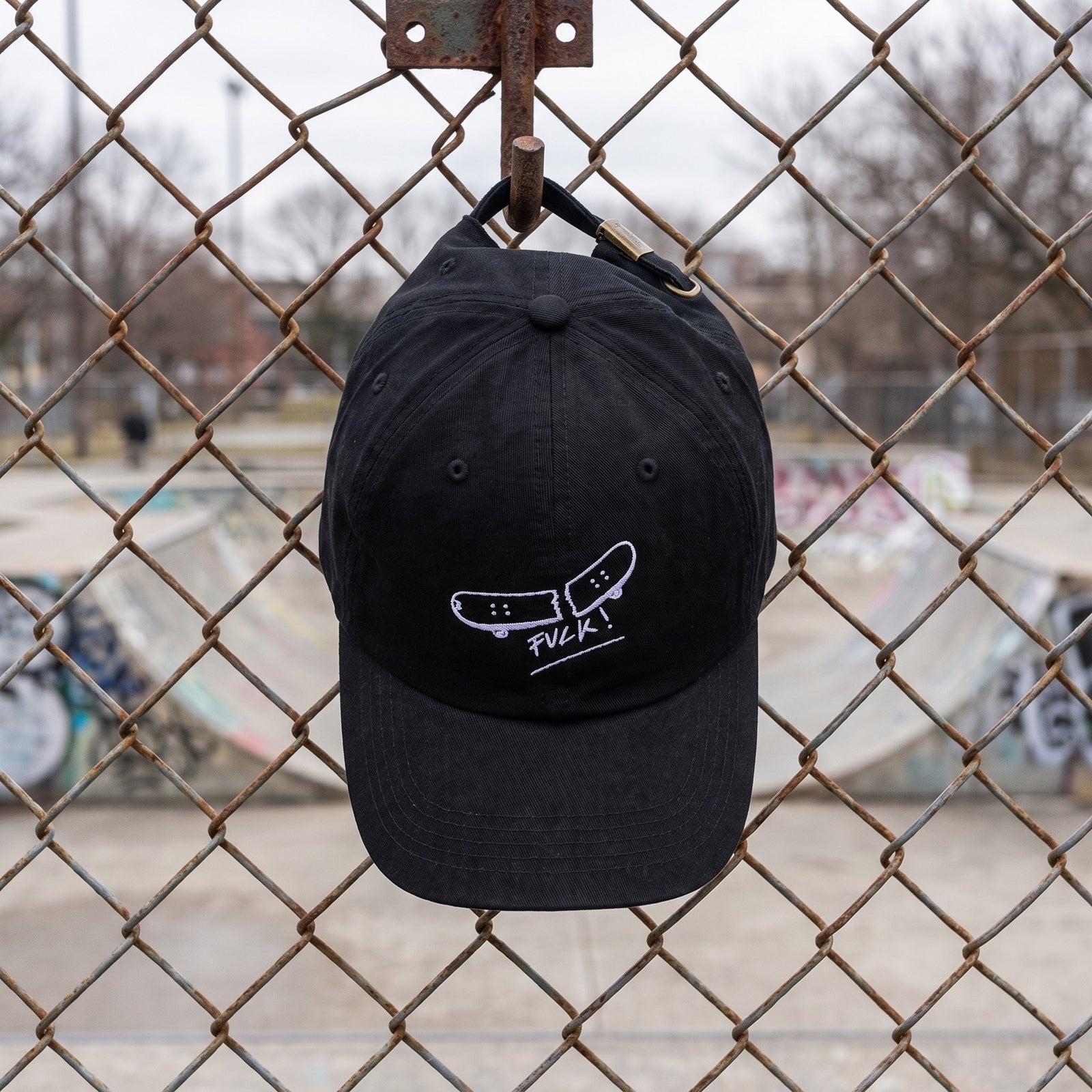 Casquette skate brodée FVCK noire suspendue à un grillage de skatepark, dad hat coton