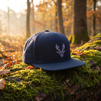 Casquette snapback bleu marine brodée d'une tête de cerf origami géométrique blanche posée sur un tronc moussu dans une forêt d'automne au lever du soleil - CHT'M Boutique