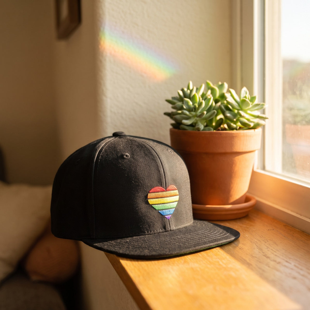 Casquette snapback noire avec un coeur arc-en-ciel brodé sur le côté posée sur le rebord d'une fenêtre ensoleillée d'un appartement à côté d'une plante en pot avec un arc-en-ciel de lumière sur le mur - CHT'M Boutique