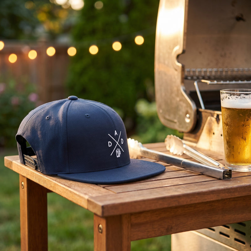 Casquette snapback bleu marine brodée du monogramme DAD blanc avec une chope de bière posée sur une desserte de barbecue dans un jardin le soir à côté de pinces et d'une bière fraîche - CHT'M Boutique