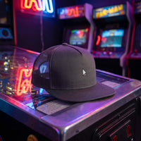 Casquette snapback gris anthracite à filet 5 panneaux brodée d'une petite flèche de souris blanche posée sur une borne d'arcade dans une salle de jeux rétro éclairée au néon - CHT'M Boutique
