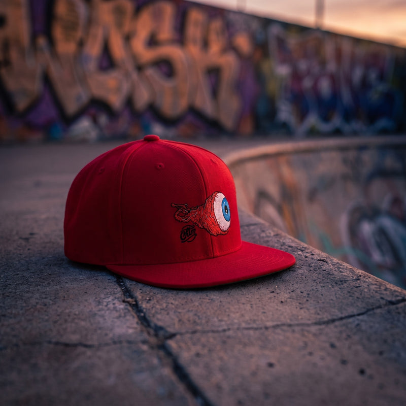 Casquette snapback rouge brodée d'un globe oculaire enflammé injecté de sang à l'iris bleu posée sur le bord en béton d'un bowl de skatepark devant un mur de graffitis au crépuscule - CHT'M Boutique