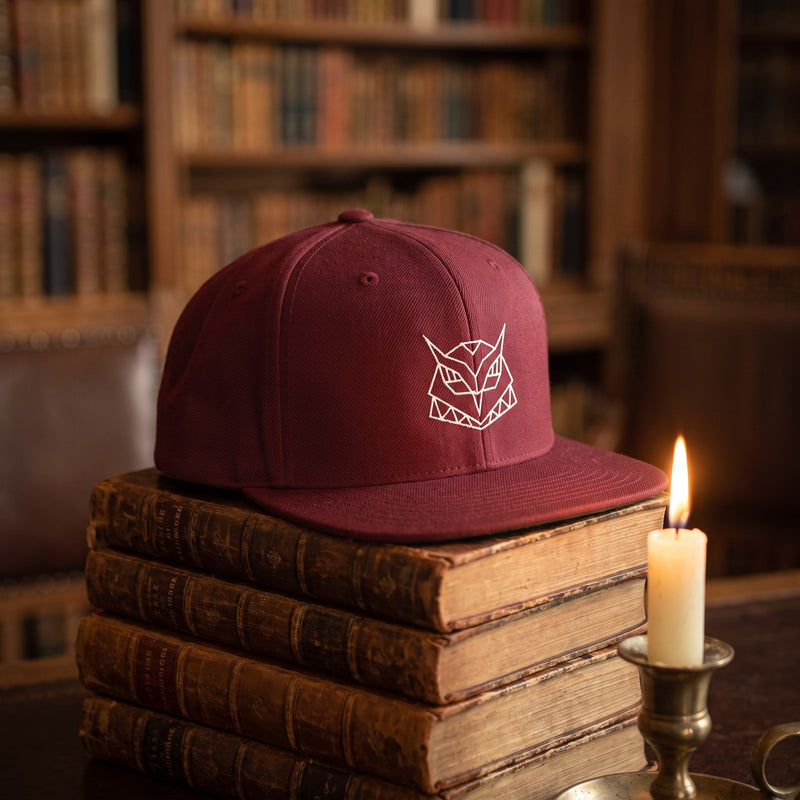 Casquette snapback bordeaux brodée d'une tête de hibou origami géométrique blanche posée sur une pile de vieux livres en cuir dans une bibliothèque à côté d'une bougie allumée - CHT'M Boutique