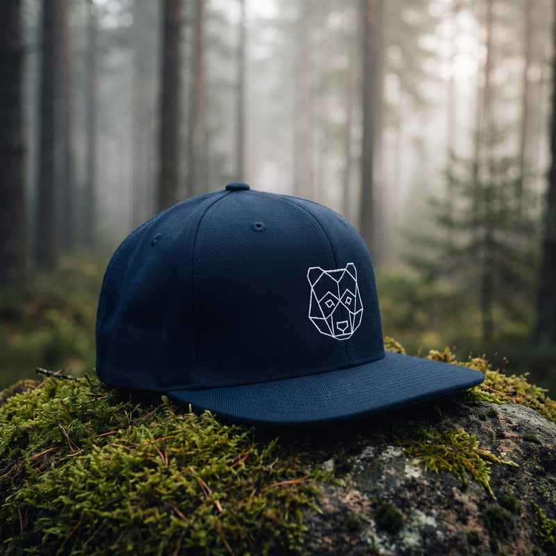 Casquette snapback bleu marine brodée d'une tête d'ours origami géométrique blanche posée sur un rocher couvert de mousse dans une forêt de pins dans la brume du matin - CHT'M Boutique