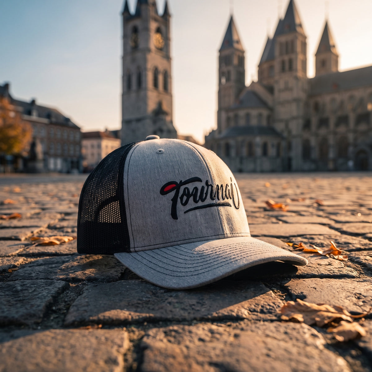 Casquette Tournai grise et noire posée sur des pavés anciens devant un beffroi et une cathédrale romane au soleil couchant