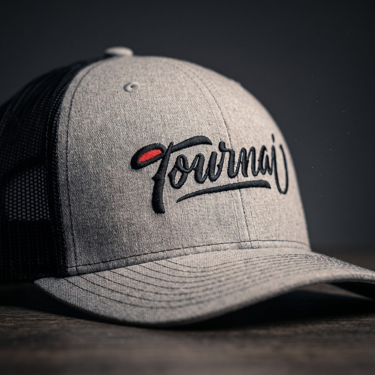 Gros plan sur la broderie script noire Tournai avec accent rouge sur une casquette trucker gris chiné