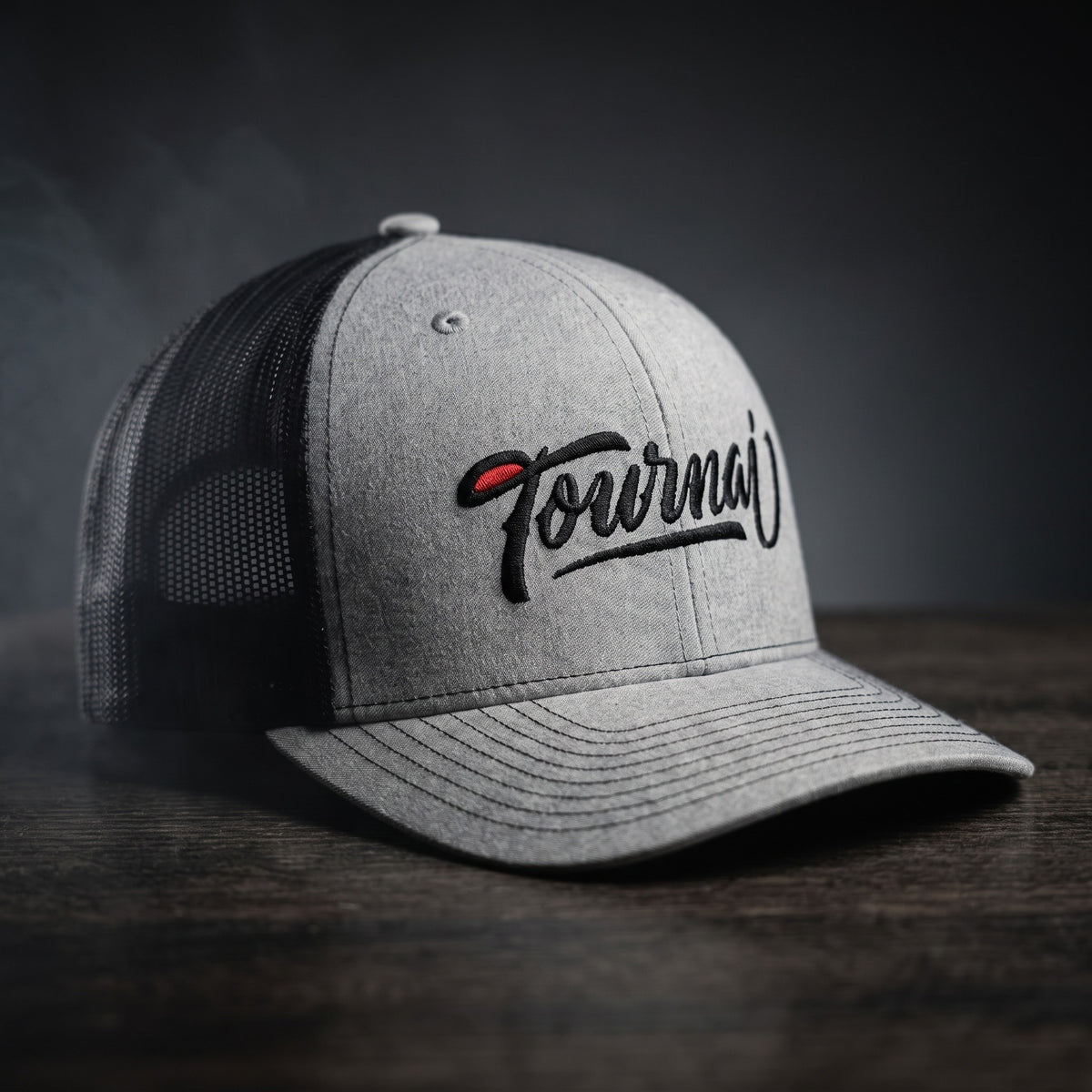 Casquette trucker gris chiné et noire brodée Tournai en script noir avec accent rouge, posée sur une table en bois sombre