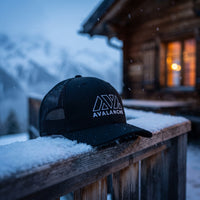 Casquette trucker AVAX Avalanche noire sur la rambarde enneigée d'un chalet de montagne au crépuscule