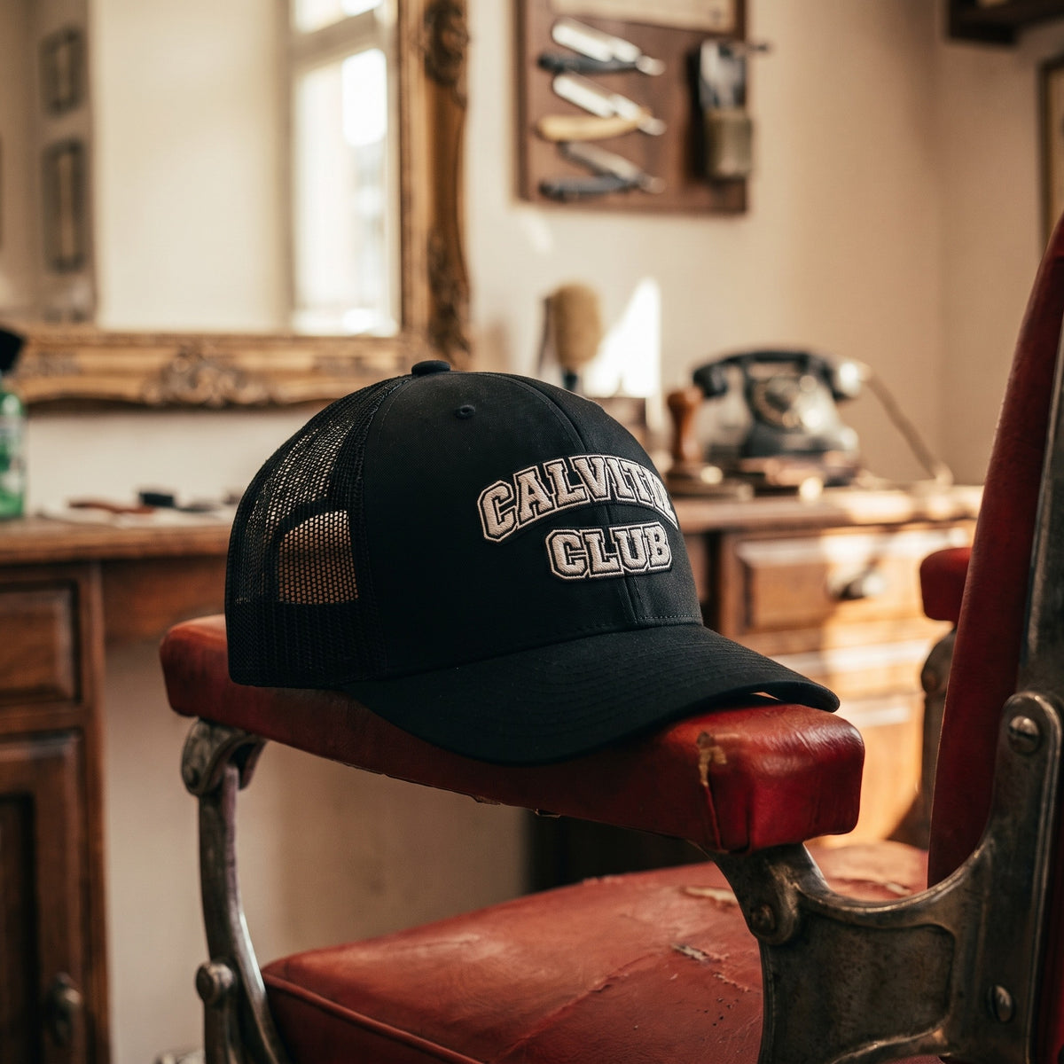 Casquette trucker CALVITIE CLUB posée sur l'accoudoir d'un fauteuil de barbier vintage dans un salon de barbier vide