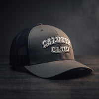 Casquette trucker brodée CALVITIE CLUB anthracite et noire posée sur un bois sombre