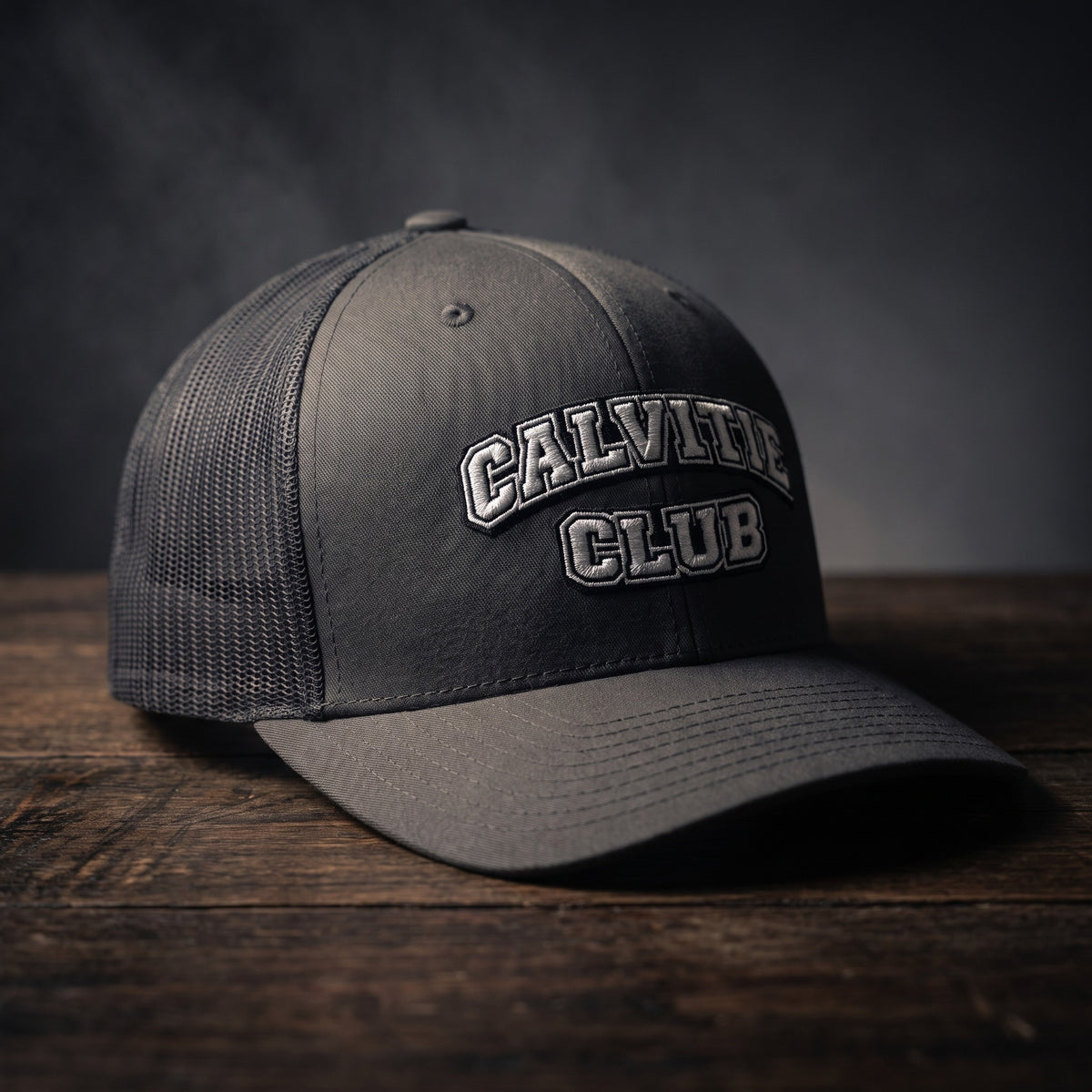 Casquette trucker brodée CALVITIE CLUB anthracite posée sur un bois sombre