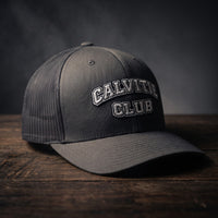 Casquette trucker brodée CALVITIE CLUB anthracite posée sur un bois sombre