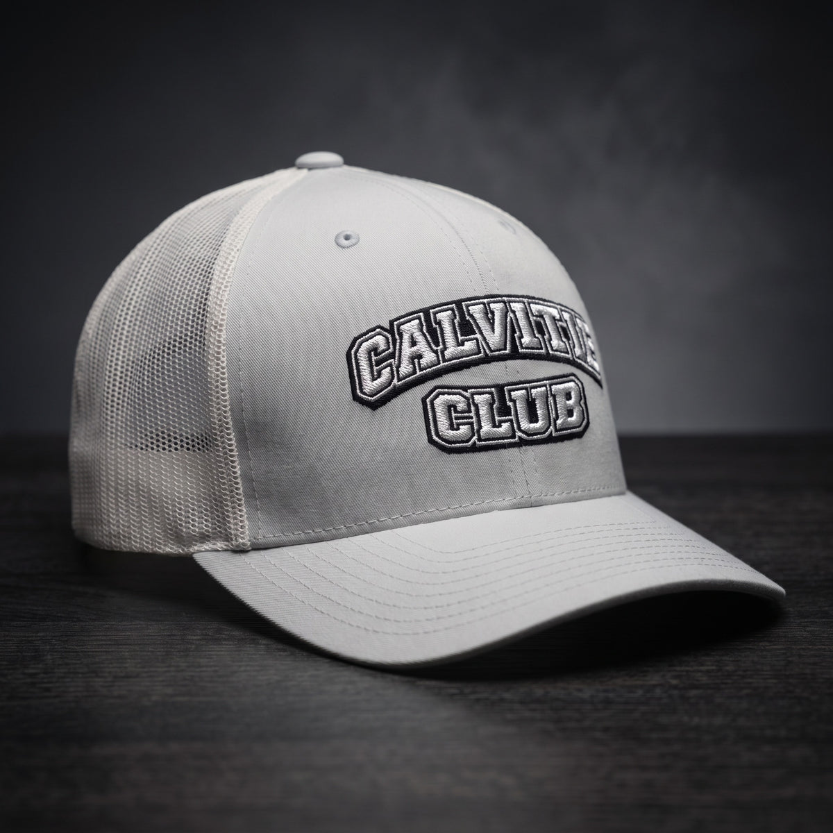 Casquette trucker brodée CALVITIE CLUB argentée posée sur un bois sombre
