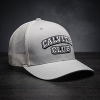 Casquette trucker brodée CALVITIE CLUB argentée posée sur un bois sombre