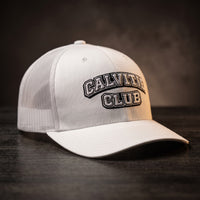Casquette trucker brodée CALVITIE CLUB blanche posée sur un bois sombre
