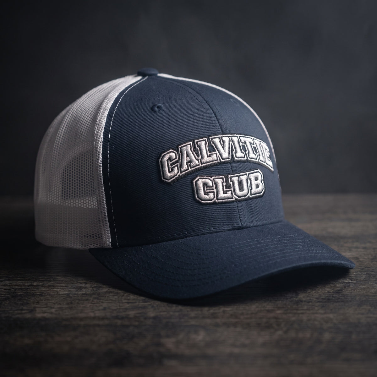 Casquette trucker brodée CALVITIE CLUB bleu marine et blanche posée sur un bois sombre