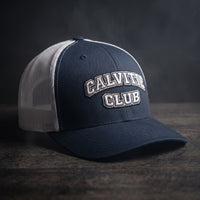 Casquette trucker brodée CALVITIE CLUB bleu marine et blanche posée sur un bois sombre