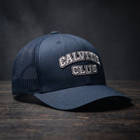 Casquette trucker brodée CALVITIE CLUB bleu marine posée sur un bois sombre