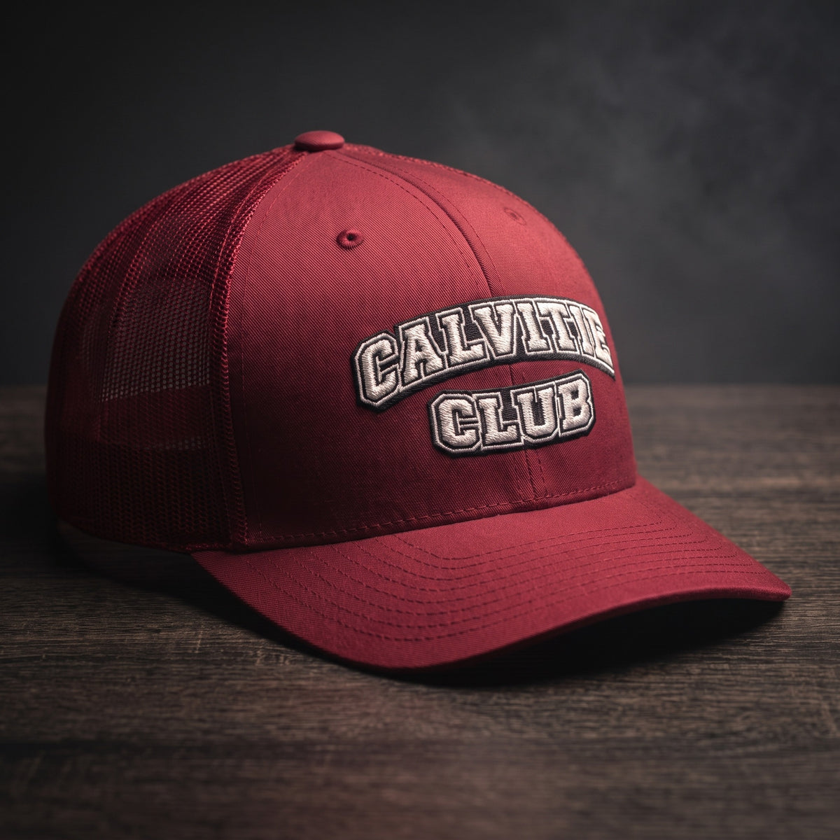 Casquette trucker brodée CALVITIE CLUB canneberge posée sur un bois sombre