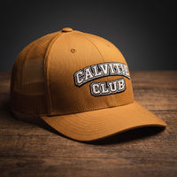 Casquette trucker brodée CALVITIE CLUB caramel posée sur un bois sombre