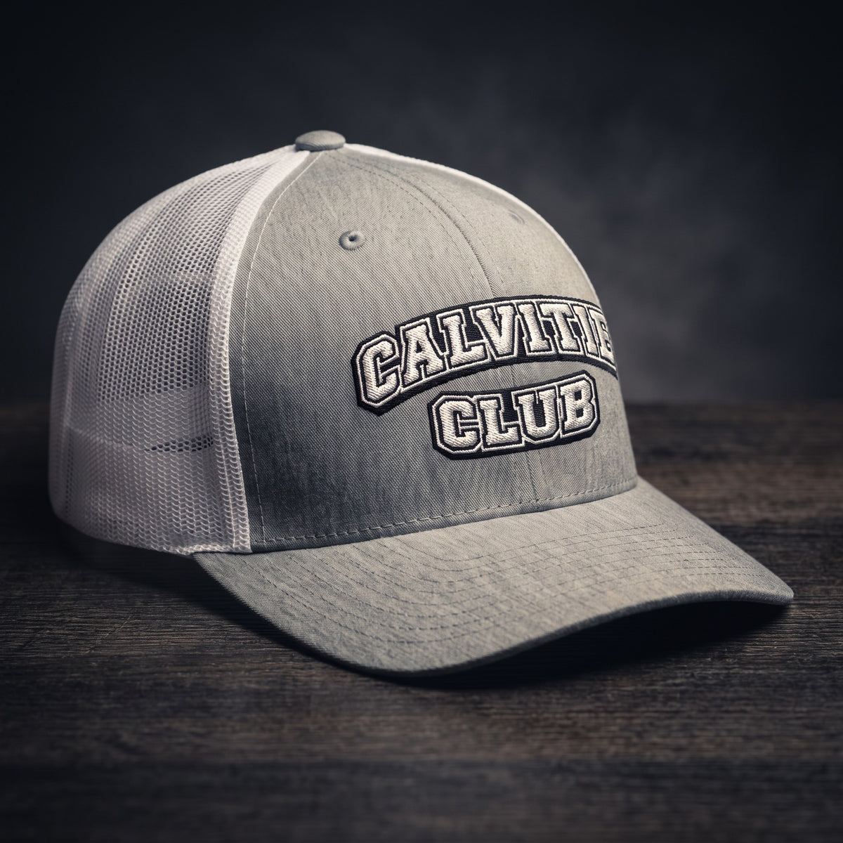 Casquette trucker brodée CALVITIE CLUB gris chiné et blanche posée sur un bois sombre
