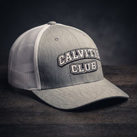 Casquette trucker brodée CALVITIE CLUB gris chiné et blanche posée sur un bois sombre