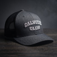 Casquette trucker brodée CALVITIE CLUB gris chiné foncé posée sur un bois sombre