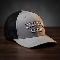Casquette trucker brodée CALVITIE CLUB gris chiné et noire posée sur un bois sombre