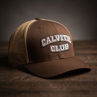 Casquette trucker brodée CALVITIE CLUB marron et kaki posée sur un bois sombre