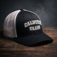 Casquette trucker brodée CALVITIE CLUB noire et blanche posée sur un bois sombre