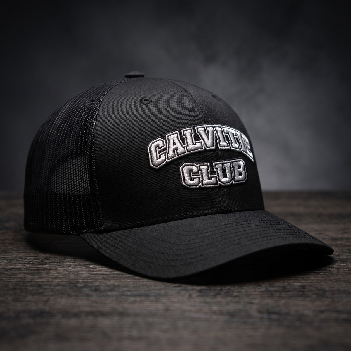 Casquette trucker brodée CALVITIE CLUB noire posée sur un bois sombre