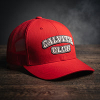 Casquette trucker brodée CALVITIE CLUB rouge posée sur un bois sombre