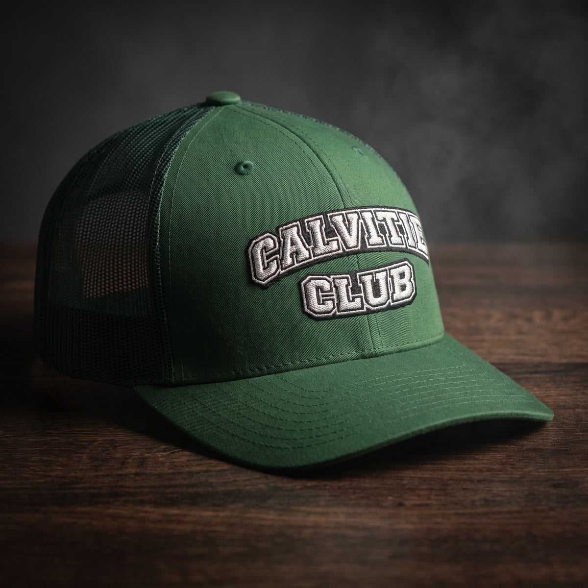 Casquette trucker brodée CALVITIE CLUB verte posée sur un bois sombre