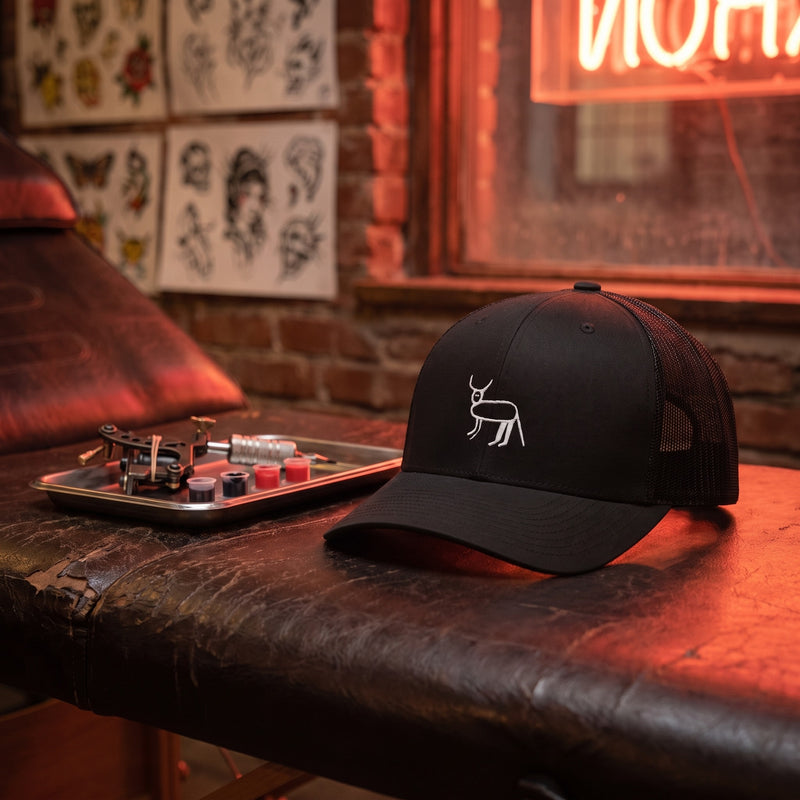 Casquette trucker noire brodée de la perruche ratée posée sur un banc en cuir dans un salon de tatouage, néon rouge et planches de flash